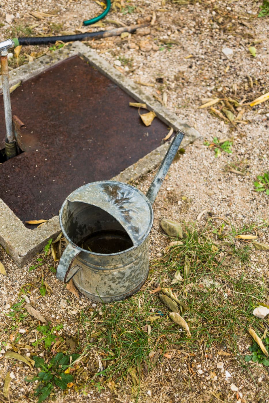 Galvanised watering can beside an open ground-level cover, with a garden hose and outdoor tap